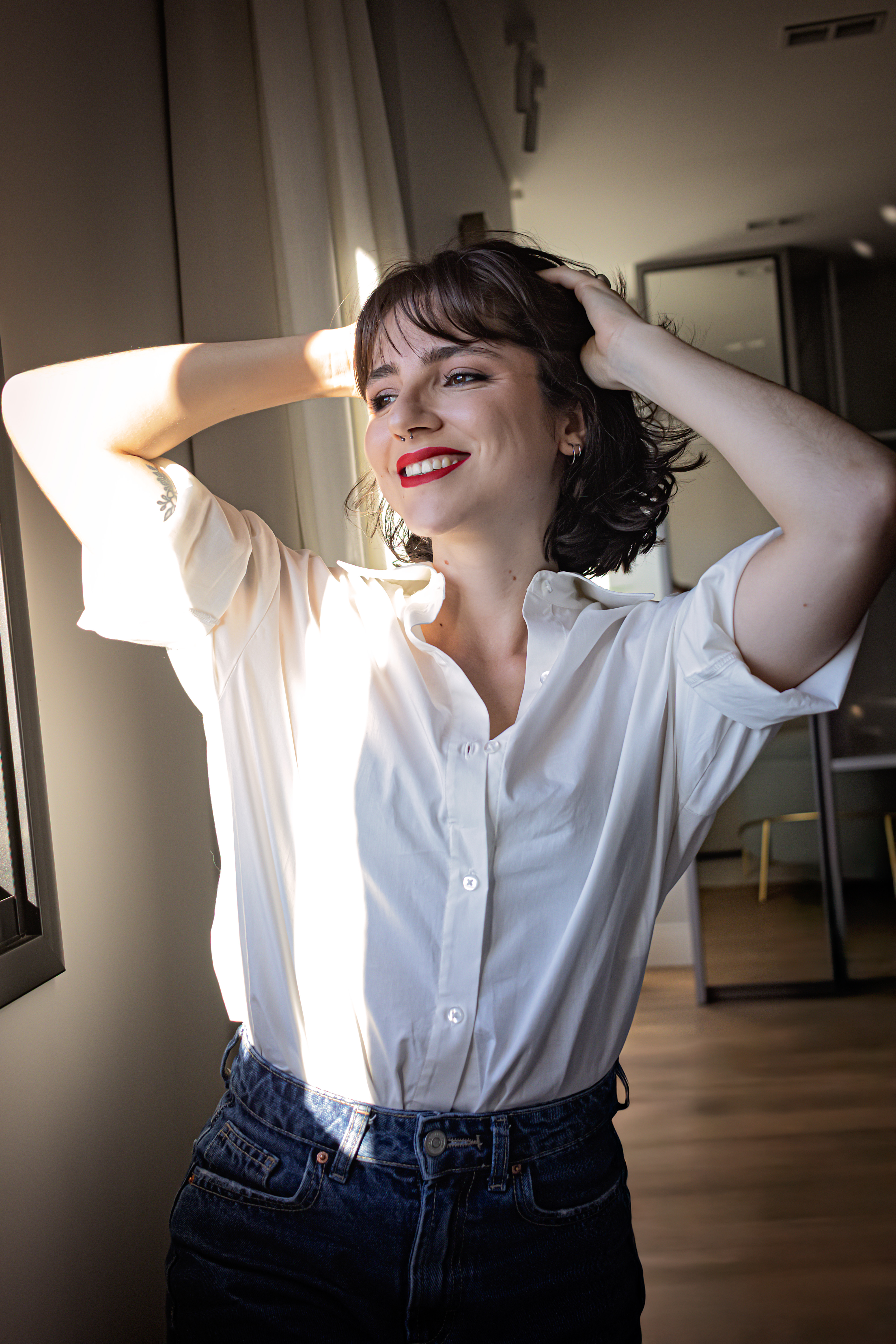 Karol Campos sorrindo junto à janela, com as mãos nos cabelos